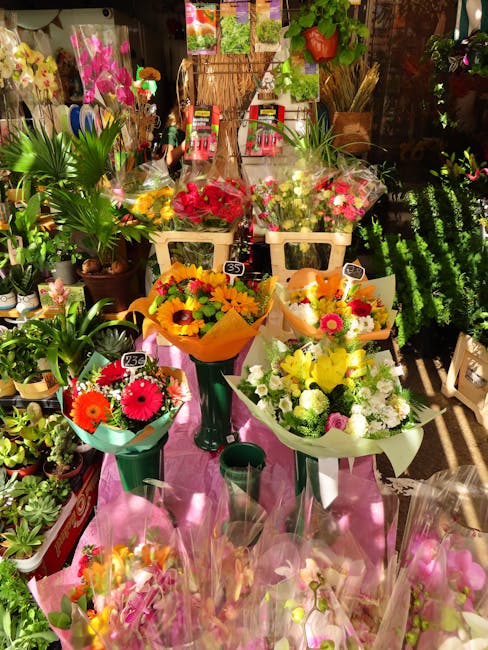 A vibrant display of fresh flower bouquets arranged on a pink cloth-covered table inside a florist shop. The bouquets feature a variety of colourful blooms, including red roses, pink gerbera daisies,